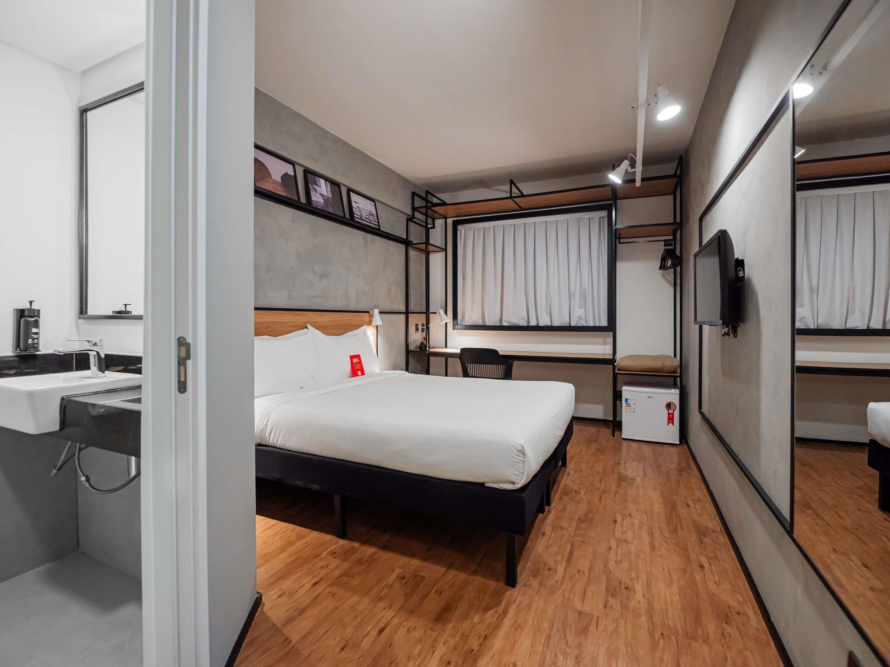 Quarto de hotel com cama, escrivaninha, cadeira, televisão e uma pequena cozinha.