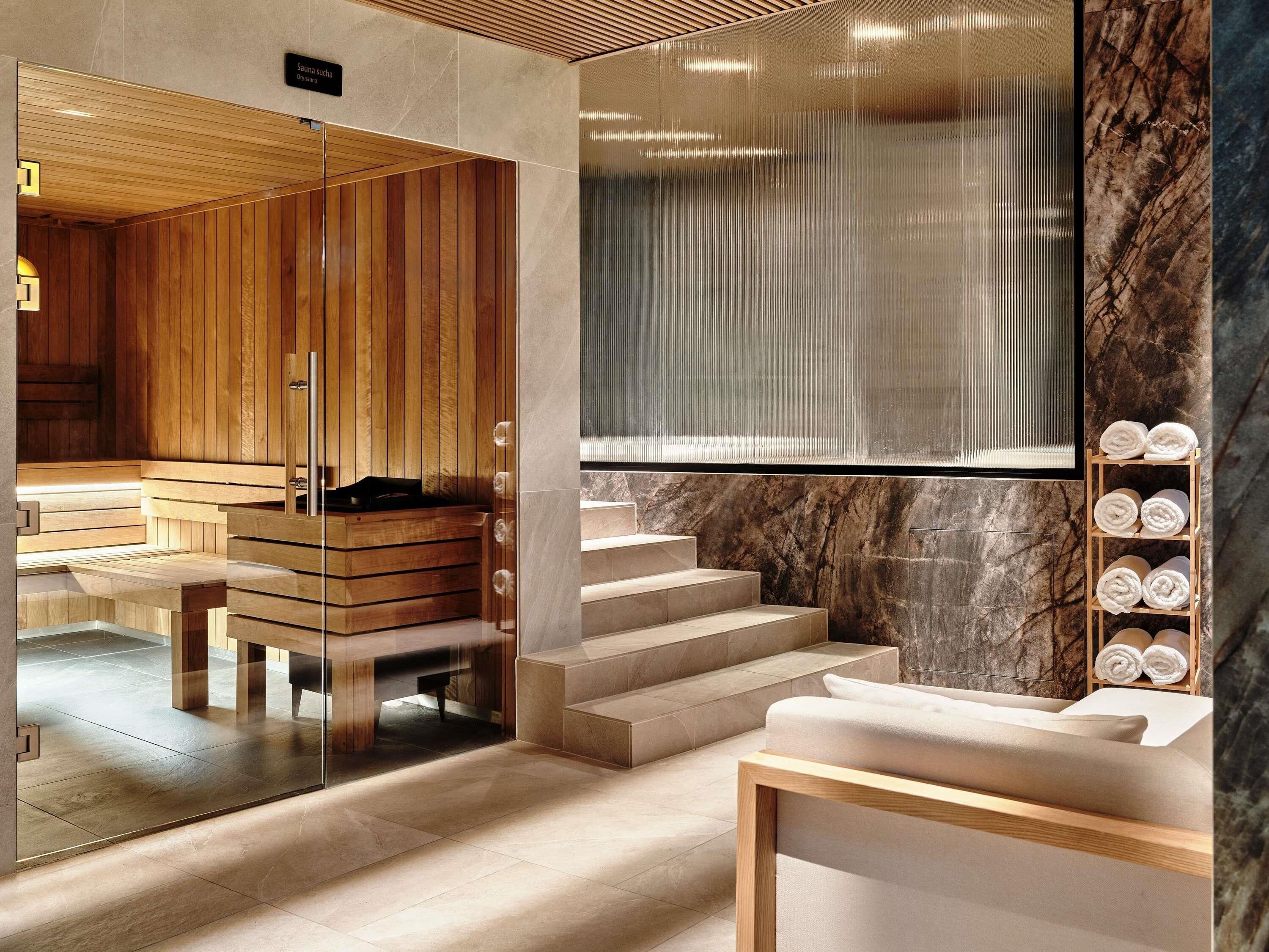 Un área de spa moderna con paneles de madera, sauna con paredes de vidrio, escalones de mármol y un estante de madera con toallas.
