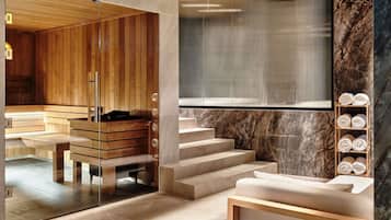 Un área de spa moderna con paneles de madera, sauna con paredes de vidrio, escalones de mármol y un estante de madera con toallas.