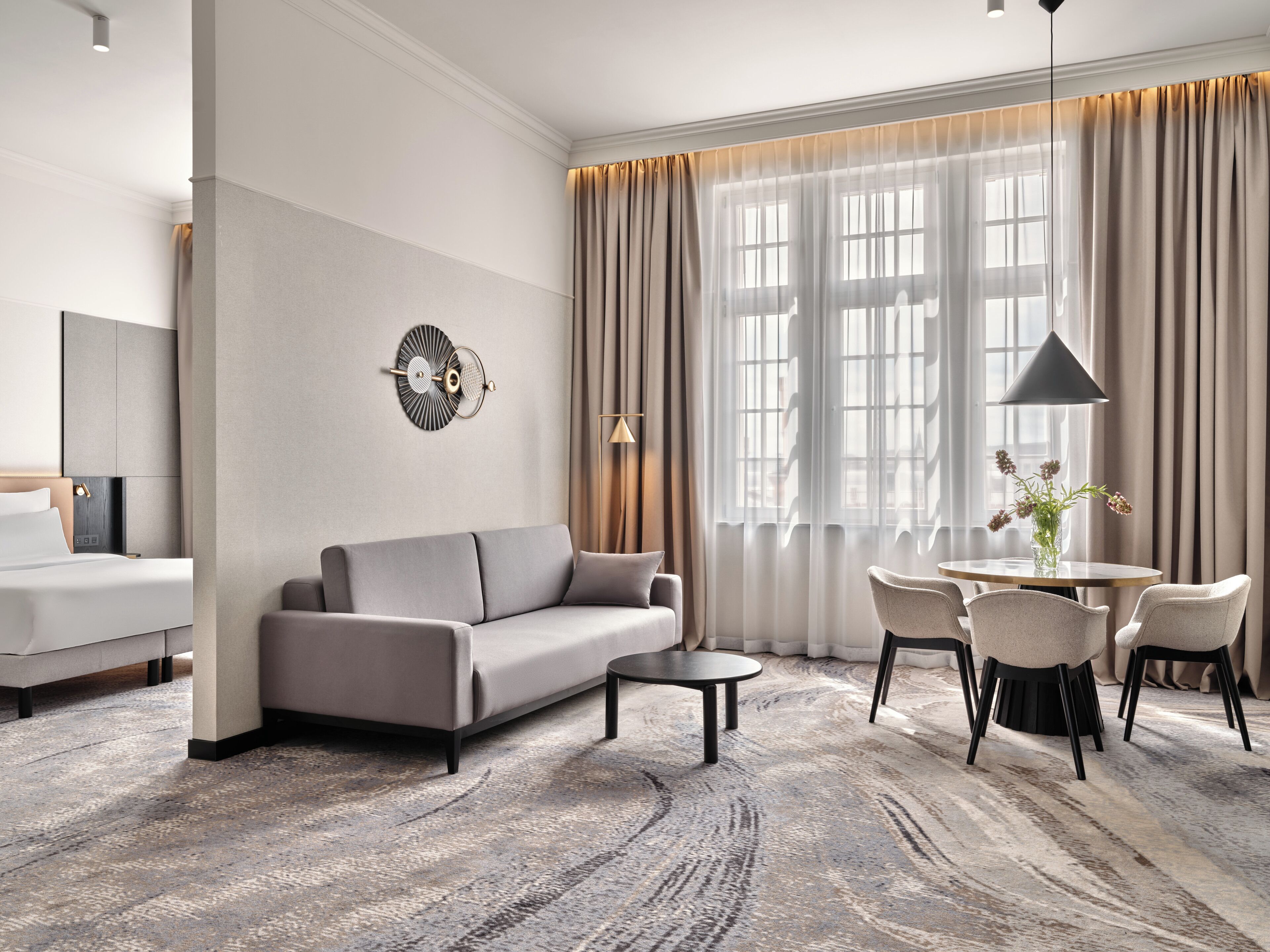 A modern hotel room with a sofa, a small table, and a chair. There is a large window with curtains, a wall-mounted lamp, and a decorative wall piece.