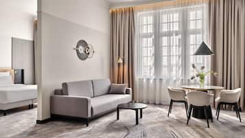 A modern hotel room with a sofa, a small table, and a chair. There is a large window with curtains, a wall-mounted lamp, and a decorative wall piece.