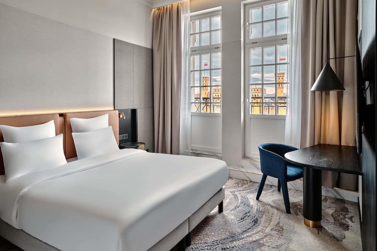 Ein Hotelzimmer mit einem großen Bett, einem Schreibtisch, einem Sessel und Blick auf Gebäude durch die Fenster.