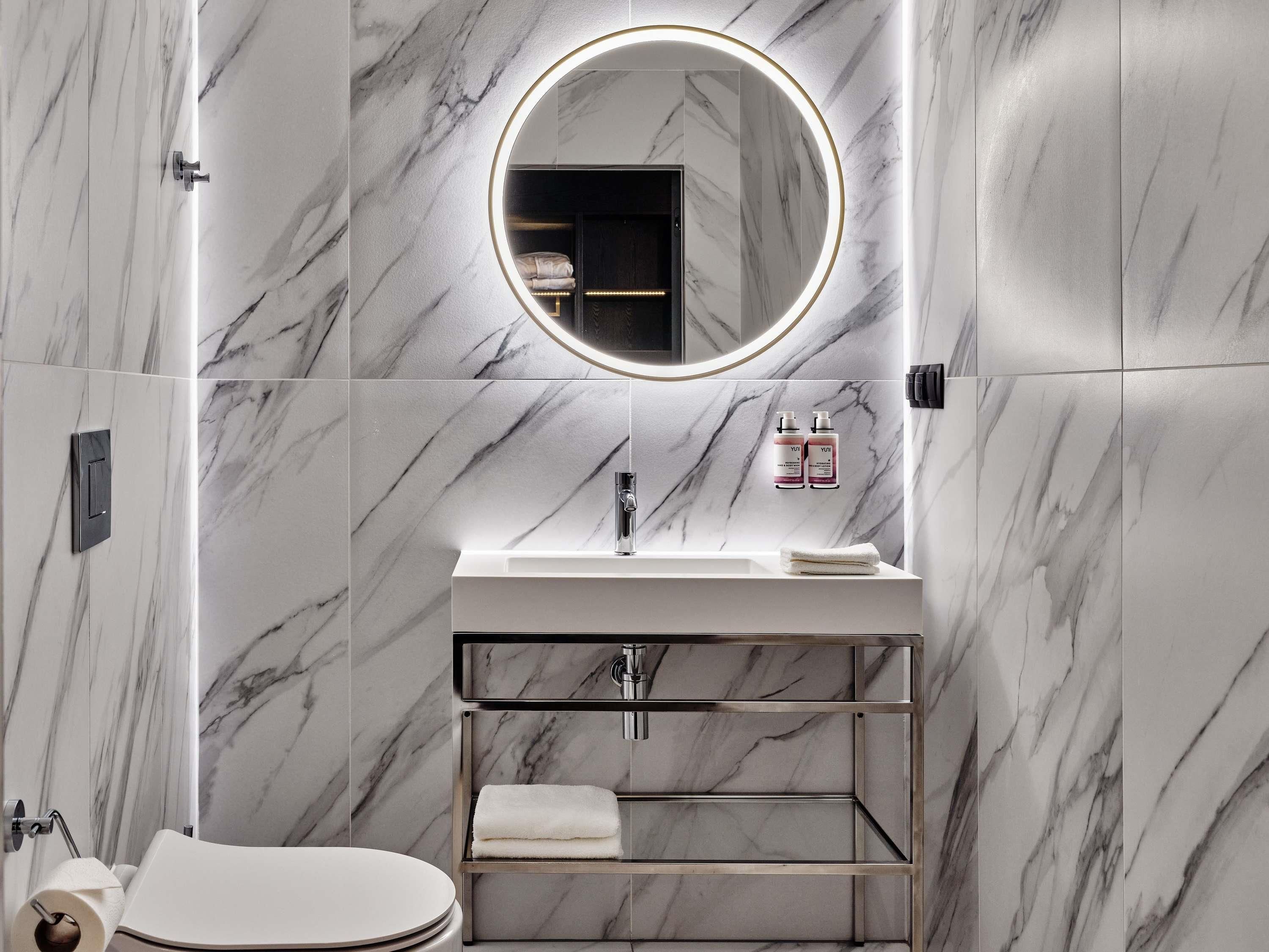 A modern bathroom with a marble wall, a round mirror, a sink, and a toilet.