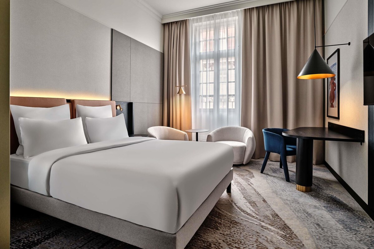A hotel room with a bed, a desk, a chair, a lamp, and a window with curtains.