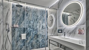 Un baño moderno con ducha envuelta en cristal, paredes de mármol, tocador con espejo y artículos de aseo.