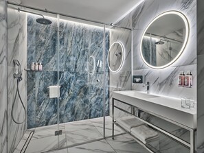 Ein modernes Badezimmer mit einer gläsernen Duschkabine, Marmorwänden, einem Waschtisch mit Spiegel und Pflegeprodukten.