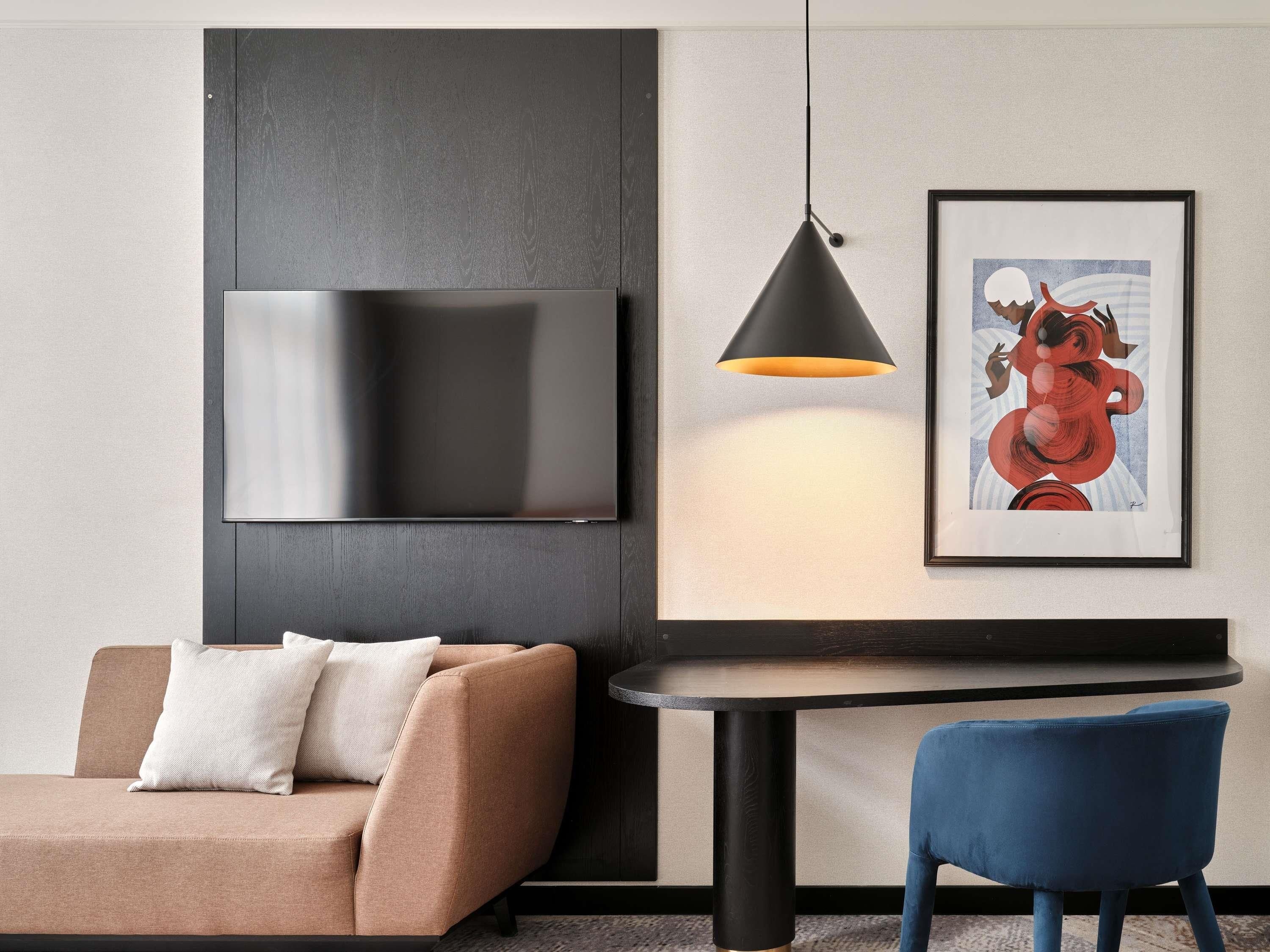 A modern room with a sofa, a table, a blue chair, a wall-mounted TV, a pendant light, and a framed artwork.