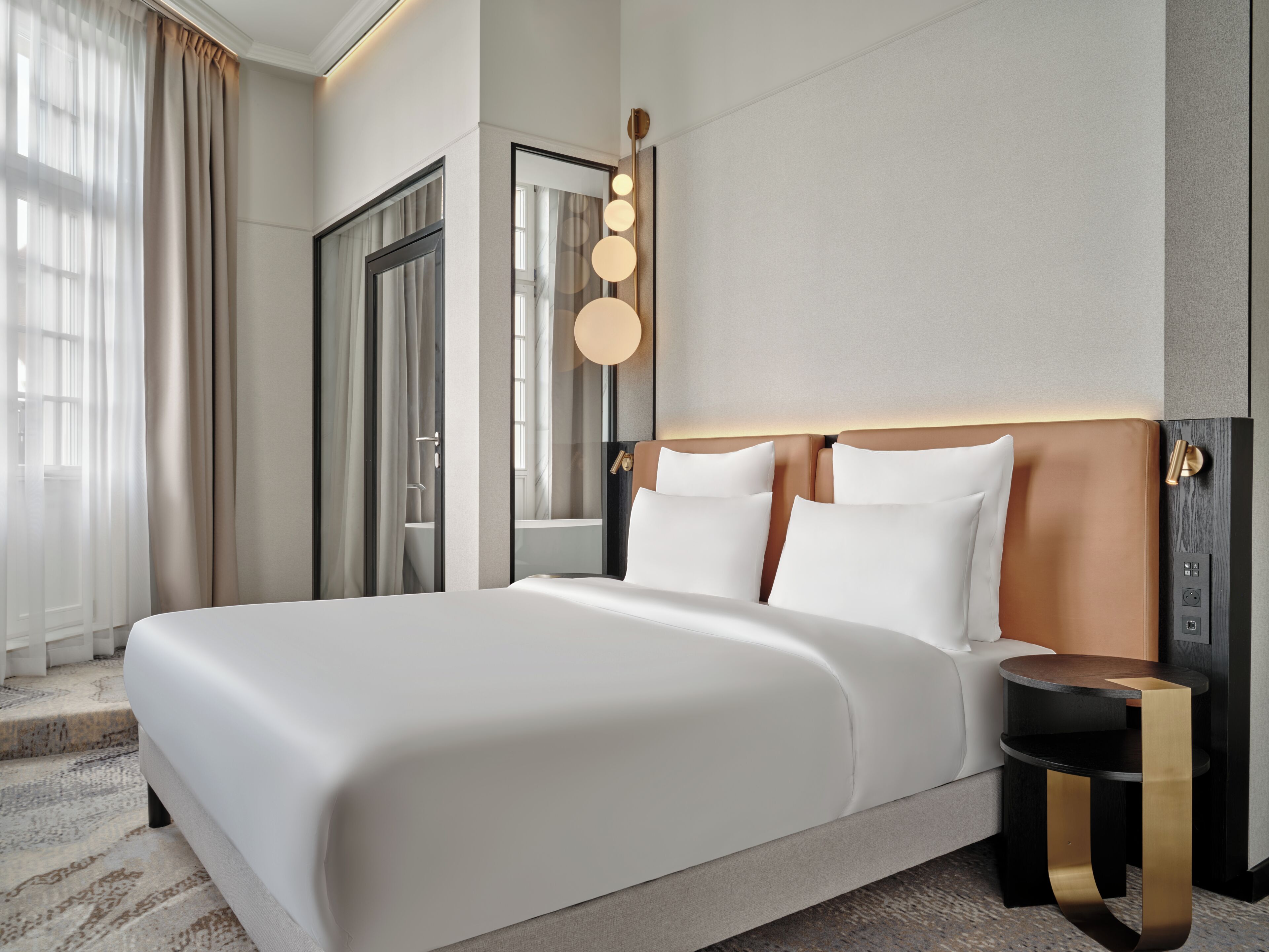 Quarto de hotel moderno com uma cama grande, mesas de cabeceira, um espelho e uma janela com cortinas.