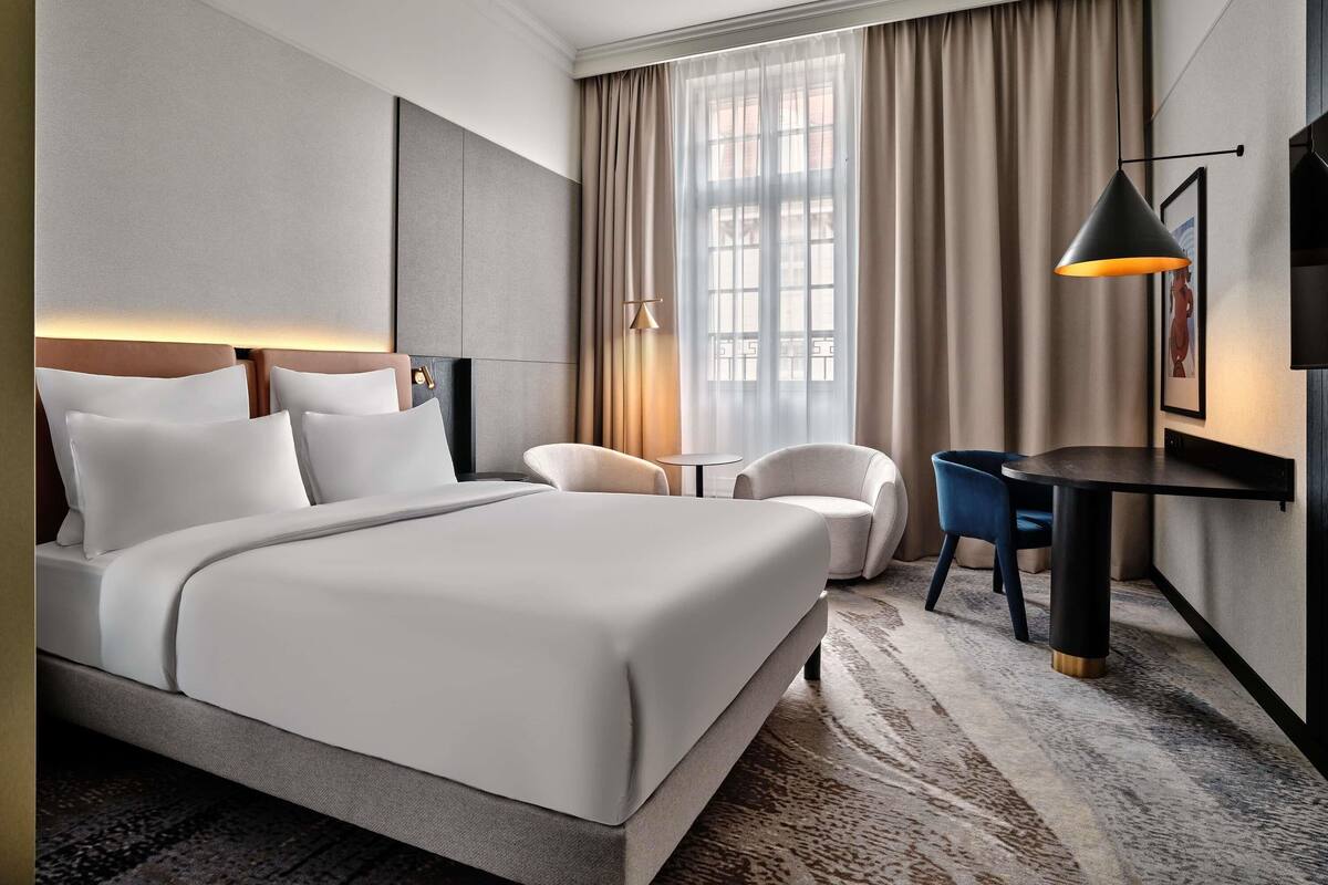 Ein Hotelzimmer mit Bett, Schreibtisch, Stuhl, Lampe und einem Fenster mit Vorhängen.