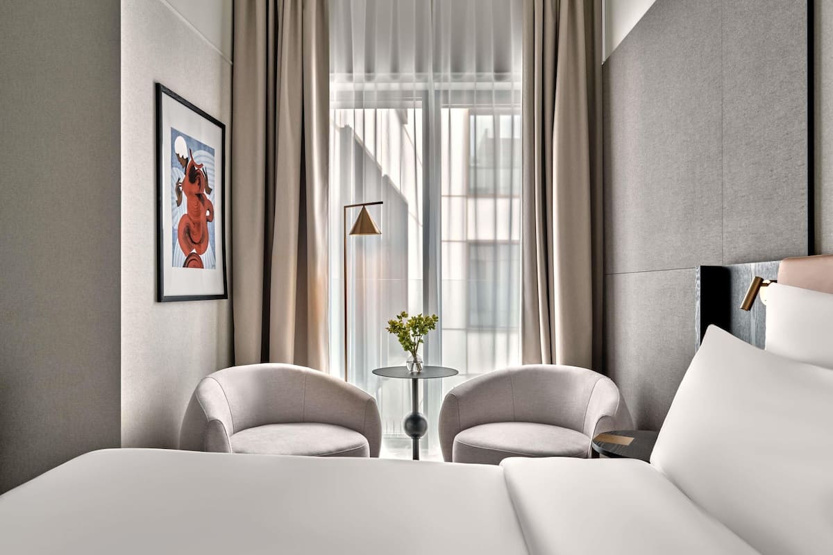 Ein modernes Hotelzimmer mit einem großen Bett, zwei Sesseln, einem kleinen Tisch mit einer Vase und einem großen Fenster mit Vorhängen.