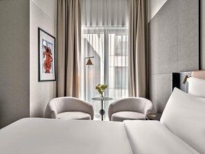 Une chambre d’hôtel moderne avec un grand lit, deux fauteuils, une petite table avec un vase et une grande fenêtre avec des rideaux.