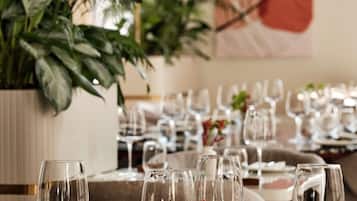 A dining area with tables set for a meal, clear glassware, and a large potted plant in the background.