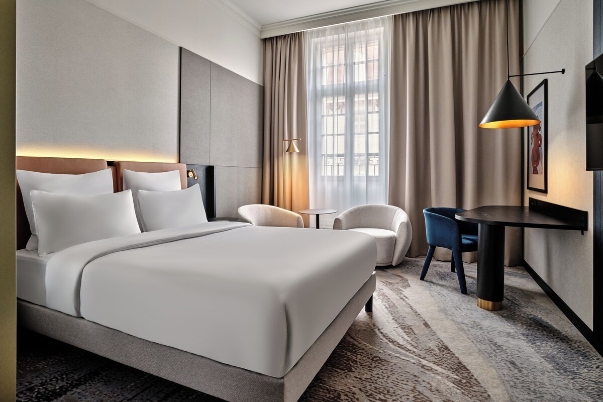 Ein Hotelzimmer mit einem großen Bett, einem Schreibtisch, einem Sessel und einem Fenster mit Vorhängen.