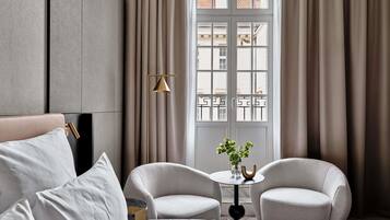 A modern hotel room with a bed, two armchairs, a small table with a vase, and a view through a window with curtains.