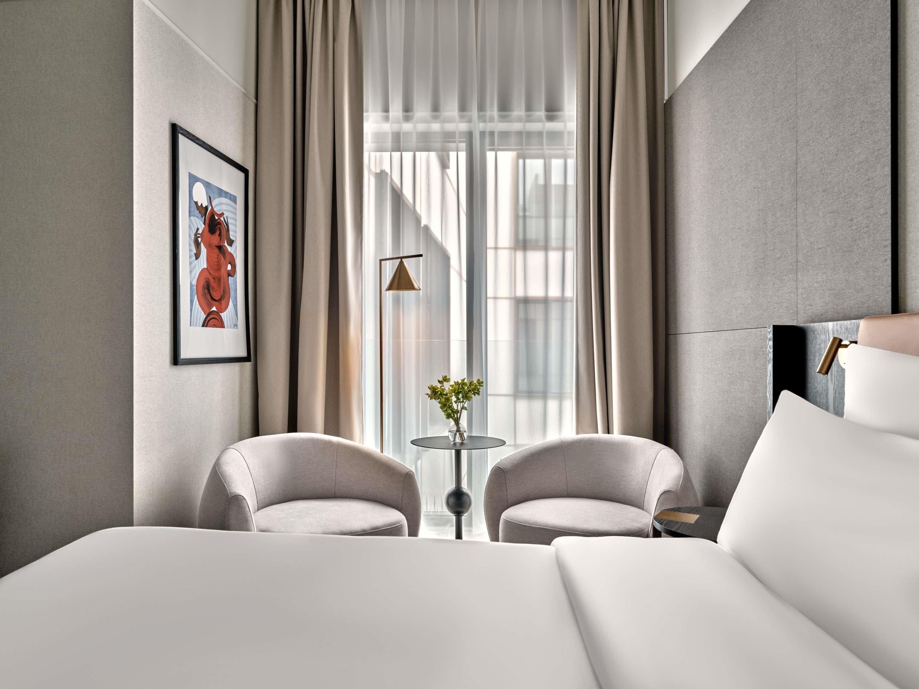 A modern hotel room with a large bed, two armchairs, a small table with a vase, and a large window with curtains.