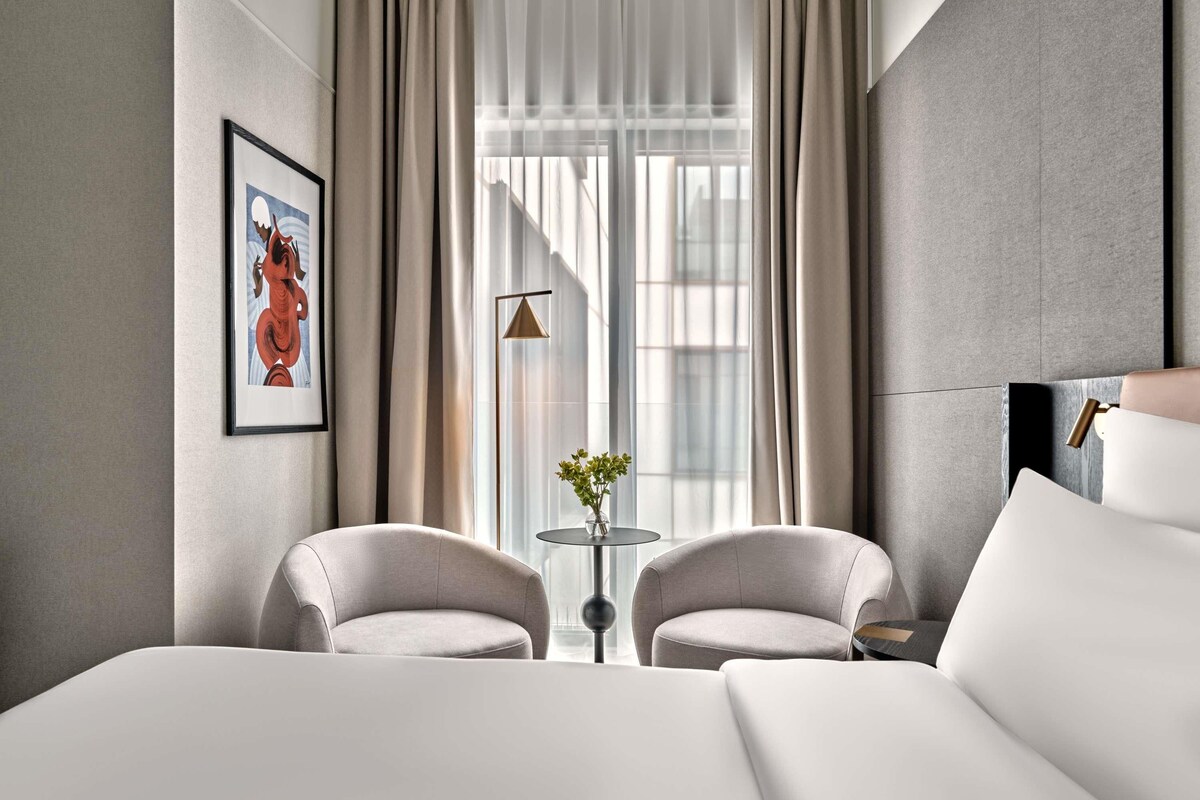 A modern hotel room with a large bed, two armchairs, a small table with a vase, and a large window with curtains.
