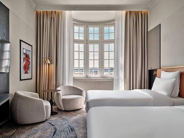 A hotel room with a bed, two armchairs, a small table, a painting, and a large window with curtains.