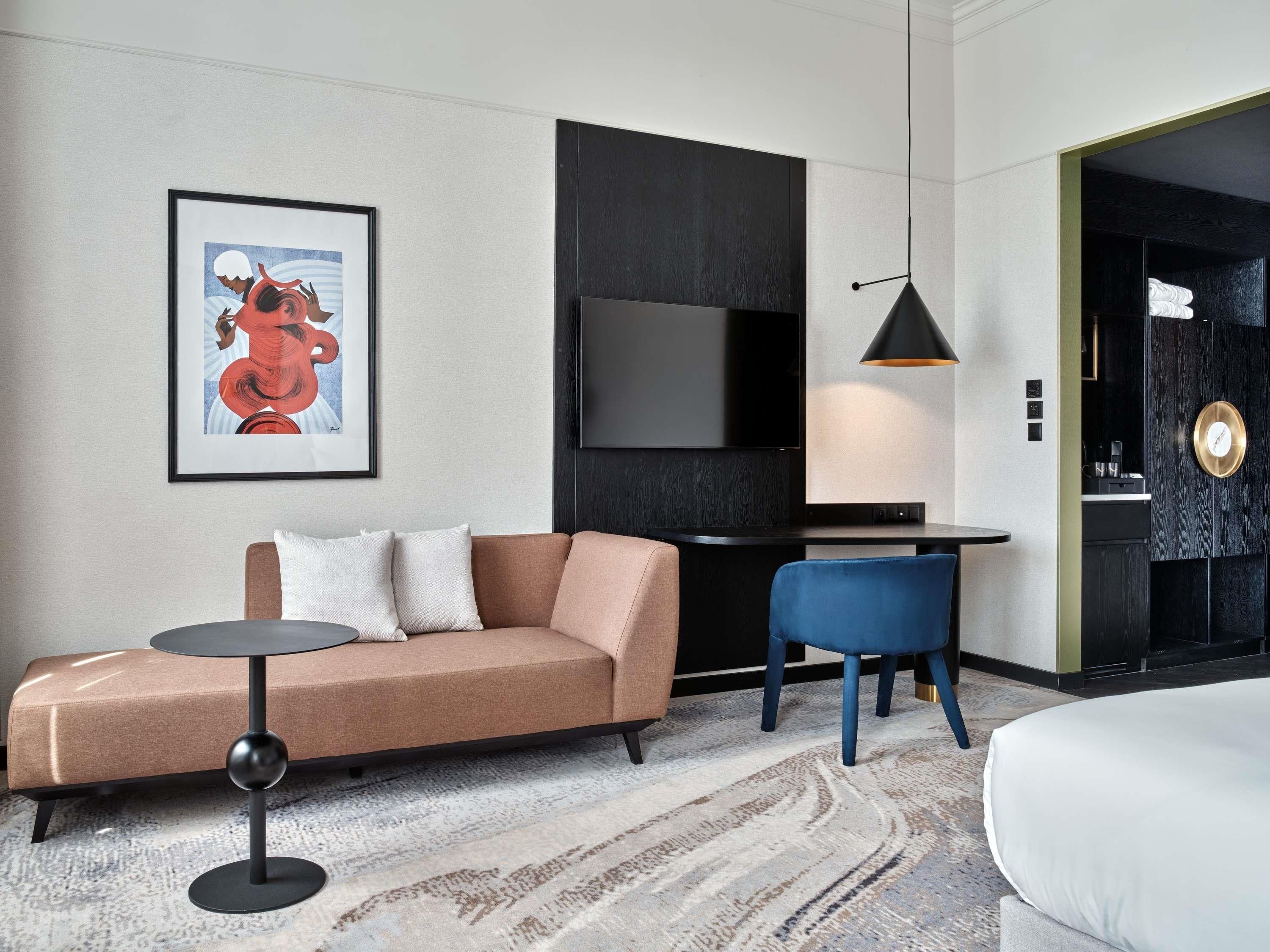 Ein modernes Hotelzimmer mit einer Couch, einem kleinen runden Tisch, einem blauen Sessel, einem an der Wand montierten Fernseher und einem Bett.