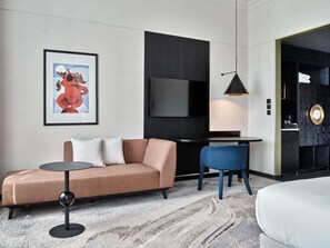 Una habitación de hotel moderna con sofá, una mesita redonda, una silla azul, televisor montado en la pared y cama.