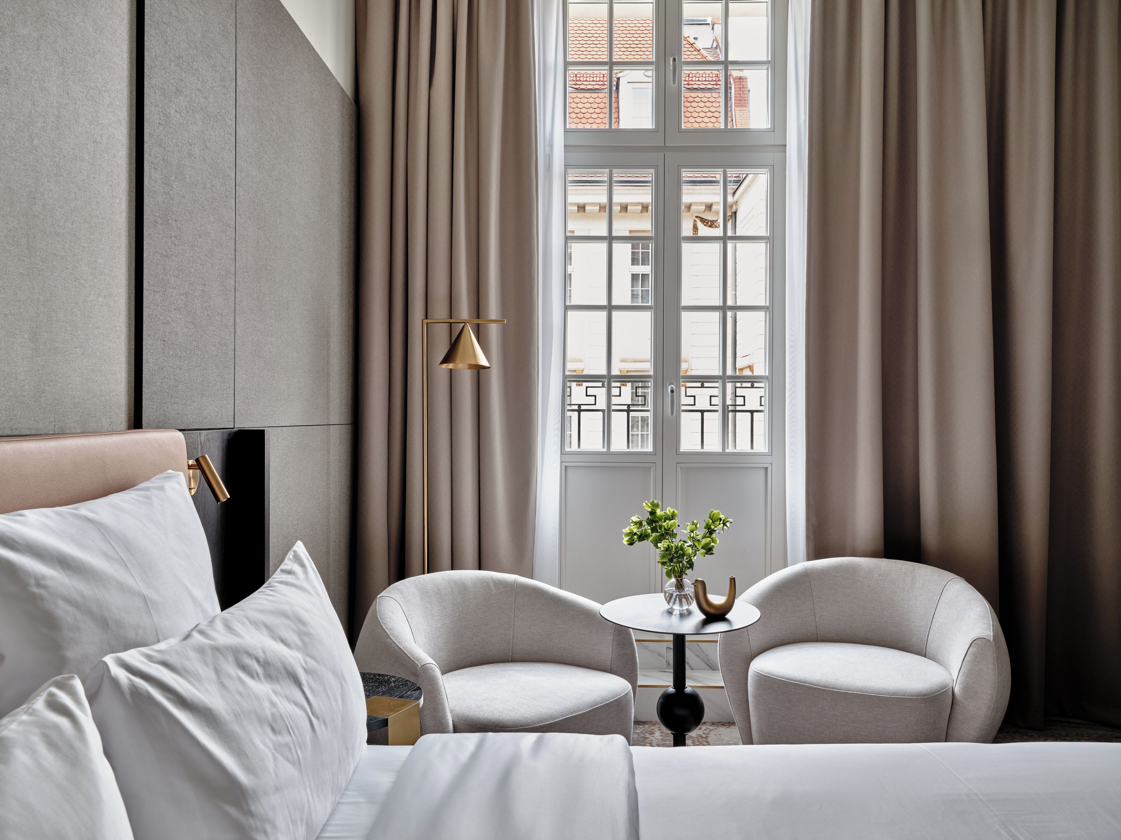 Een hotelkamer met een bed, twee fauteuils, een tafeltje met een vaas, en een uitzicht op gebouwen door een raam met gordijnen.