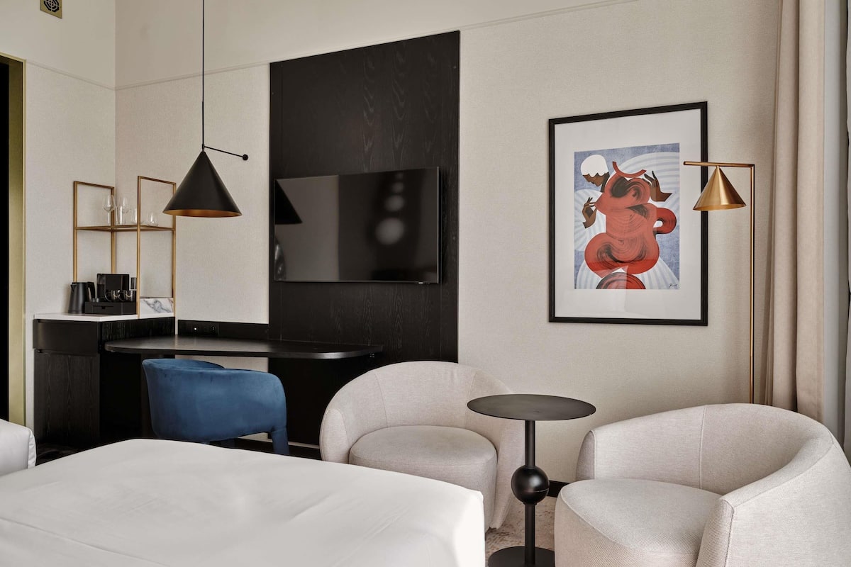 Une chambre d’hôtel comprenant un lit, deux fauteuils, une petite table ronde, un bureau avec une chaise, une télévision et deux lampes suspendues.