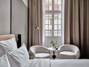 Moderni hotellihuone, jossa on sänky, kaksi nojatuolia, pieni pöytä maljakolla ja ikkunasta avautuva näkymä, jonka edessä on verhot.
