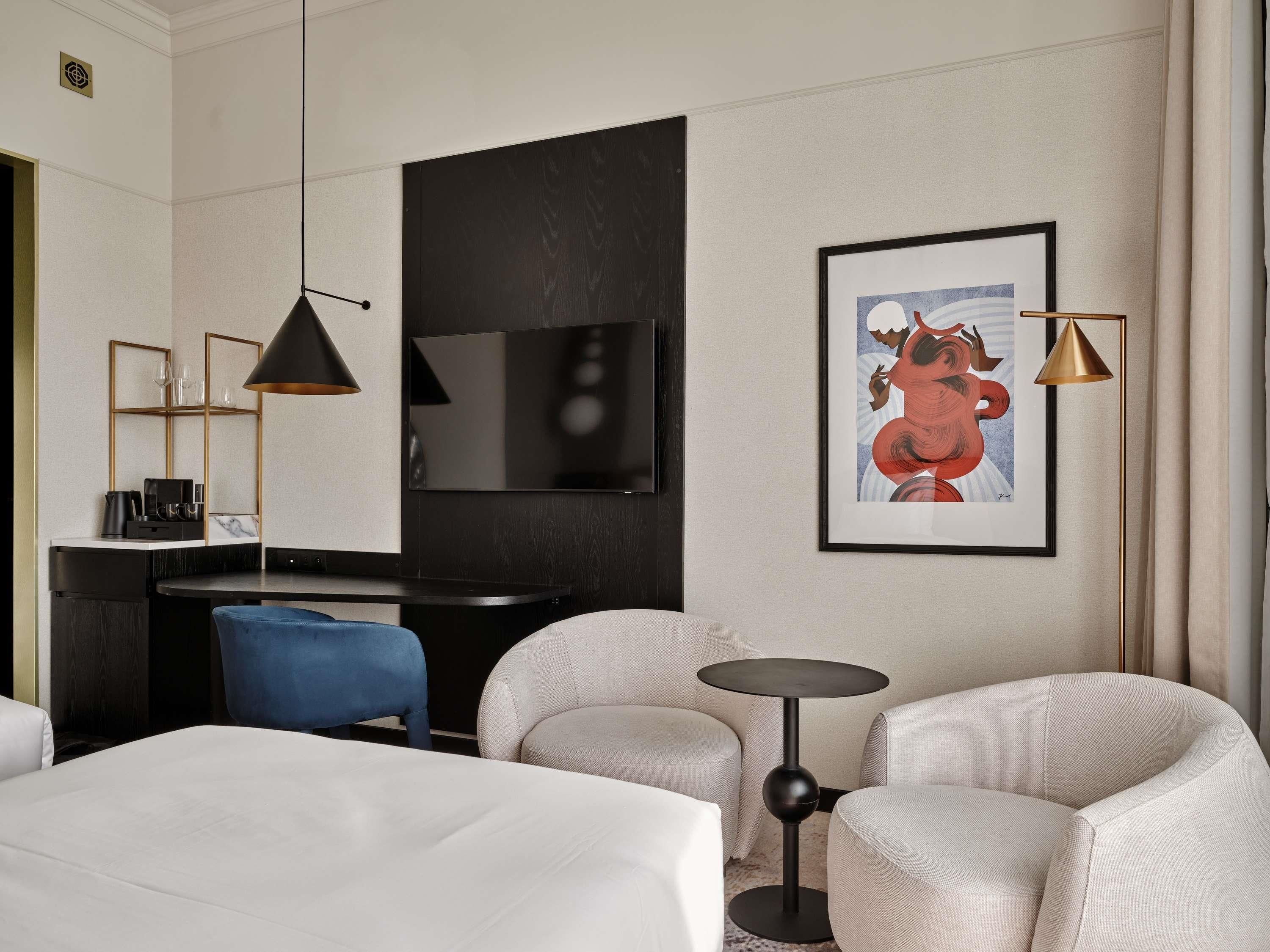 A hotel room with a bed, two armchairs, a small round table, a desk with a chair, a TV, and two pendant lights.
