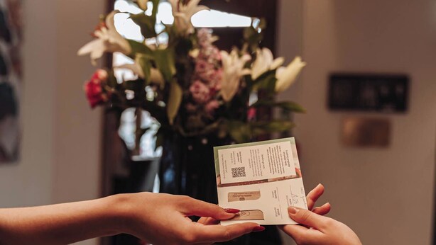 Una persona sosteniendo una tarjeta con un código QR, de pie frente a una lámpara y un arreglo floral.