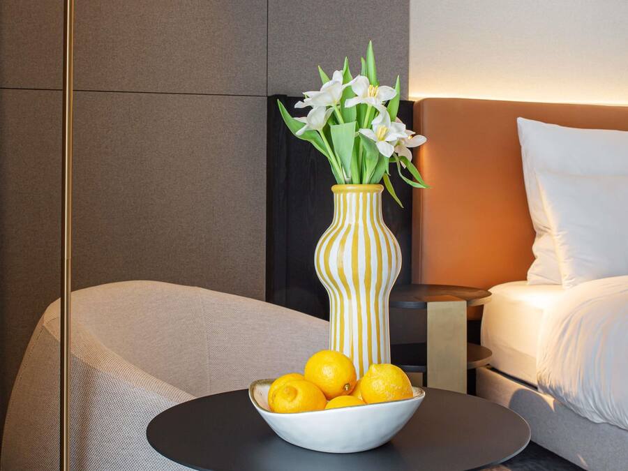 Een moderne hotelkamer met een bed, een nachtkastje met een bloevas en een schaal met citroenen op een ronde tafel.