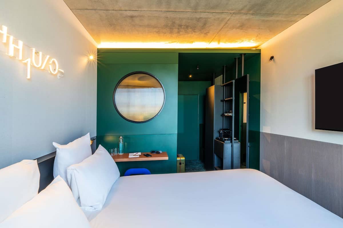 A modern hotel room with a large bed, a round mirror, a desk with a chair, and a wardrobe.