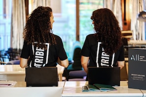 Dos personas sentadas a una mesa con portátiles, con camisetas negras que tienen la palabra "TRIBE" impresa en la parte posterior.