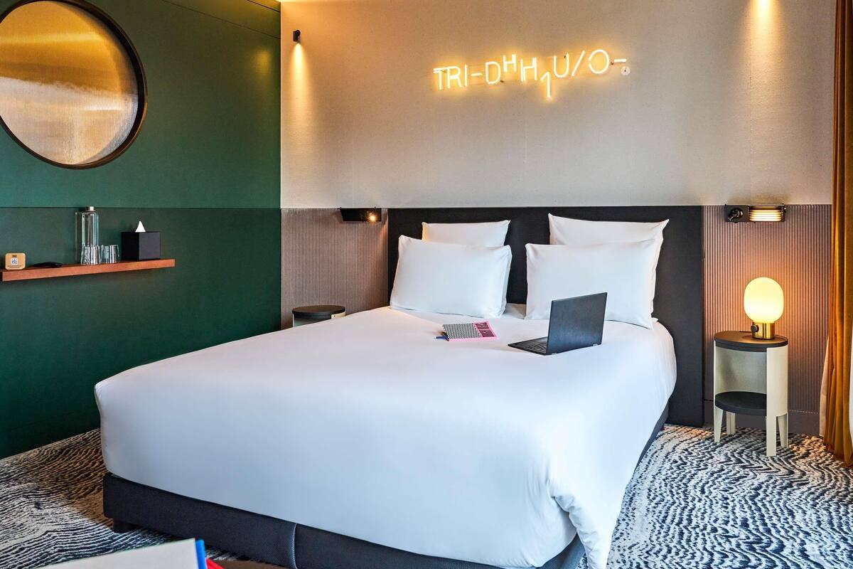 A modern hotel room with a neatly made bed, a bedside lamp, and a neon sign on the wall.