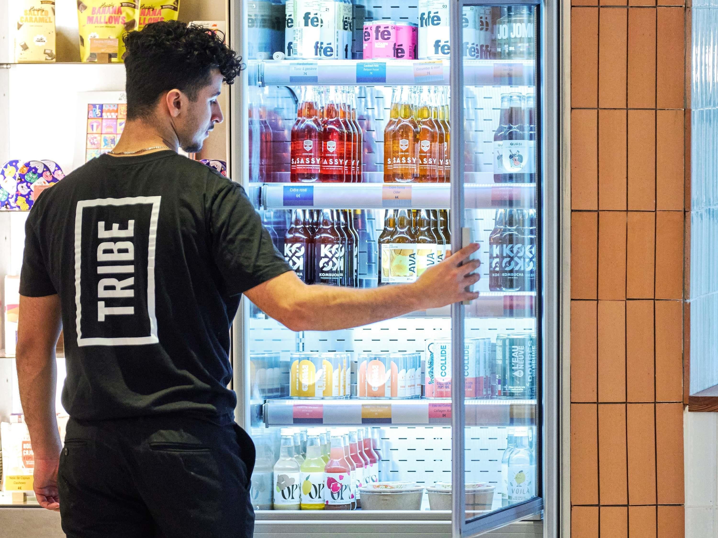 Een persoon in een zwart T-shirt met 'TRIBE' op de rug, die in een gekoelde vitrine reikt met verschillende drankjes en flessen.