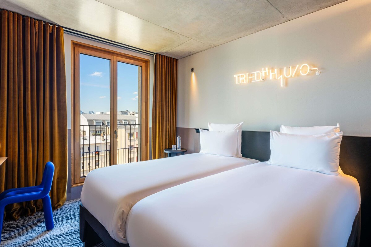 A hotel room with two beds, a view of the city through large windows, and a neon sign on the wall.