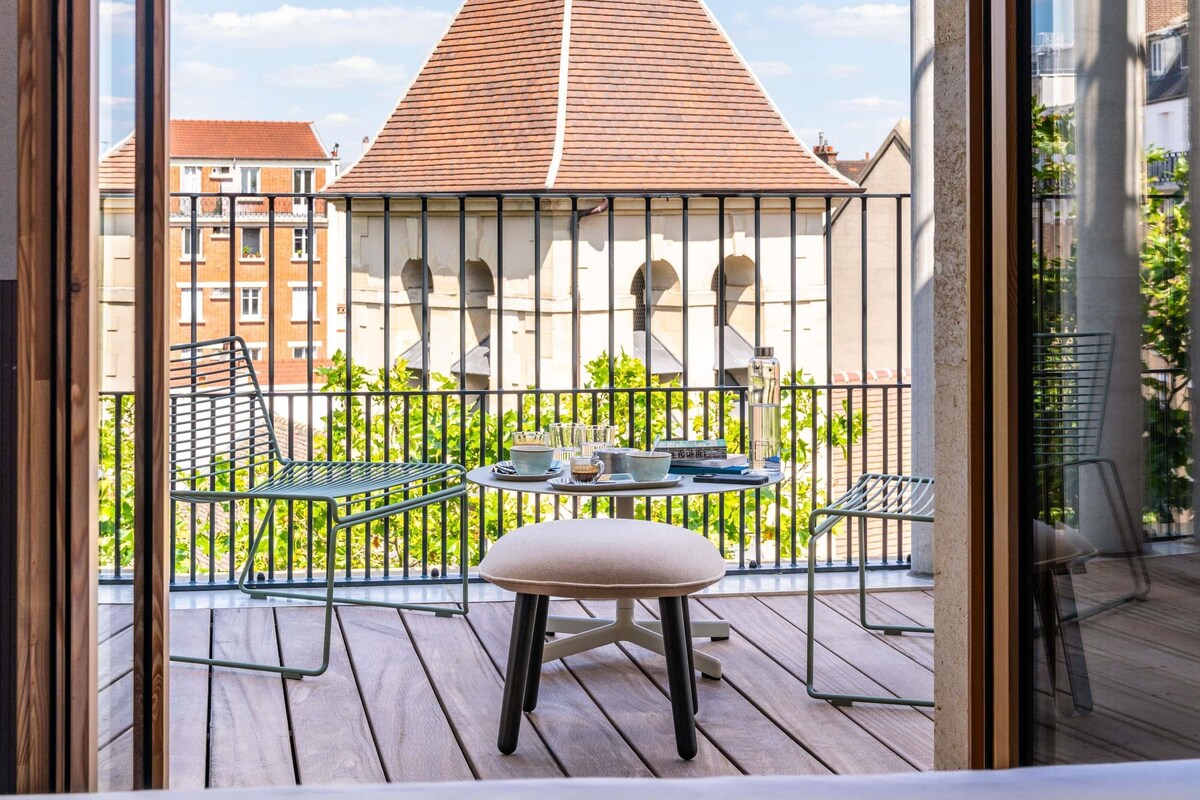 A balcony with a table, chairs, and a view of a building with a red roof.