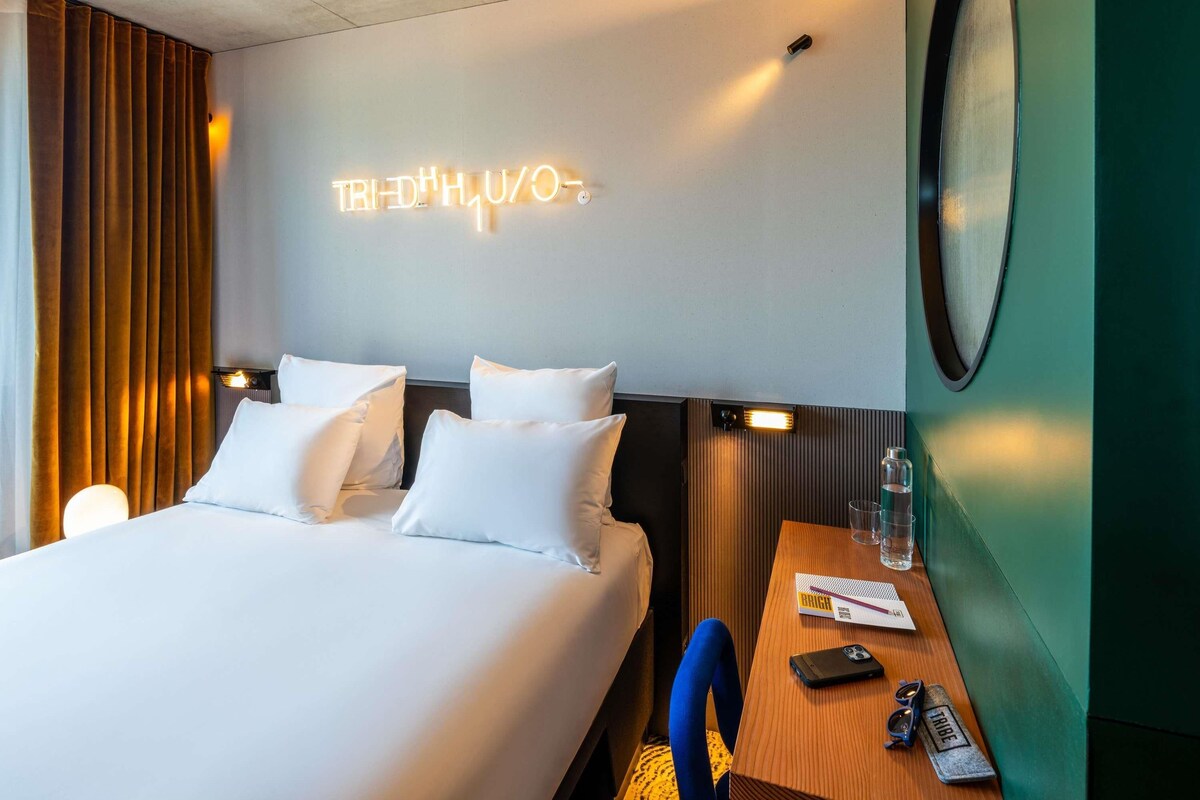 A hotel room with a bed, two pillows, a bedside table with a bottle, glasses, and a phone.