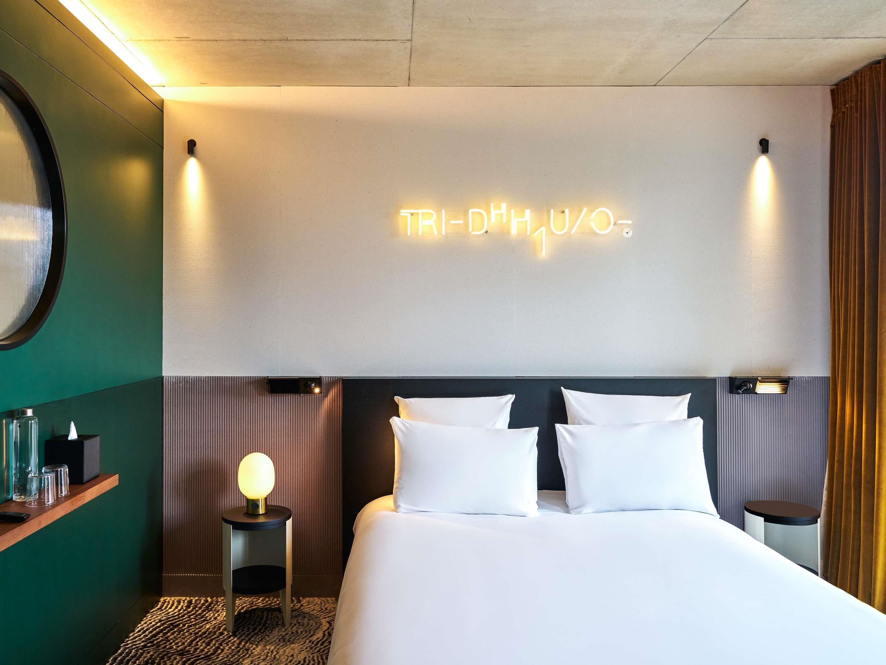 A hotel room with a bed, bedside tables, a wall-mounted light, and a neon sign.