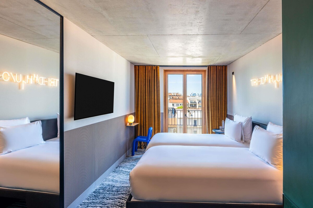 A hotel room with two beds, a TV, a small table with a lamp, and a view of the city through large windows.