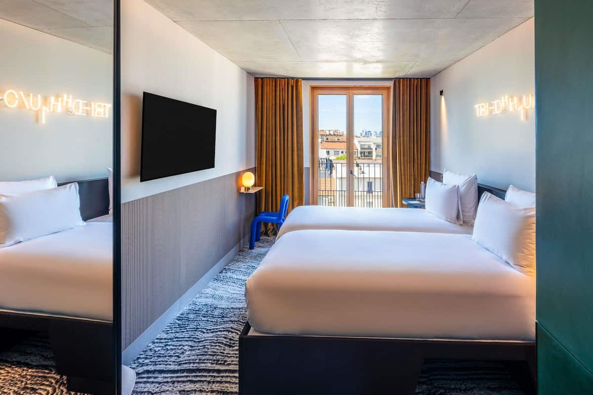 A hotel room with two beds, a TV, a small table with a lamp, and a view of the city through large windows.