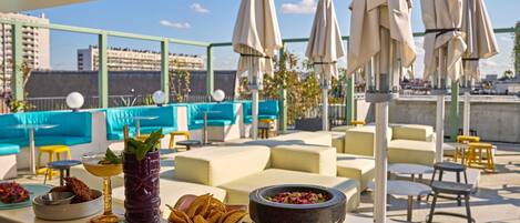 A rooftop bar with outdoor seating, a bar counter with snacks and drinks, and umbrellas providing shade.