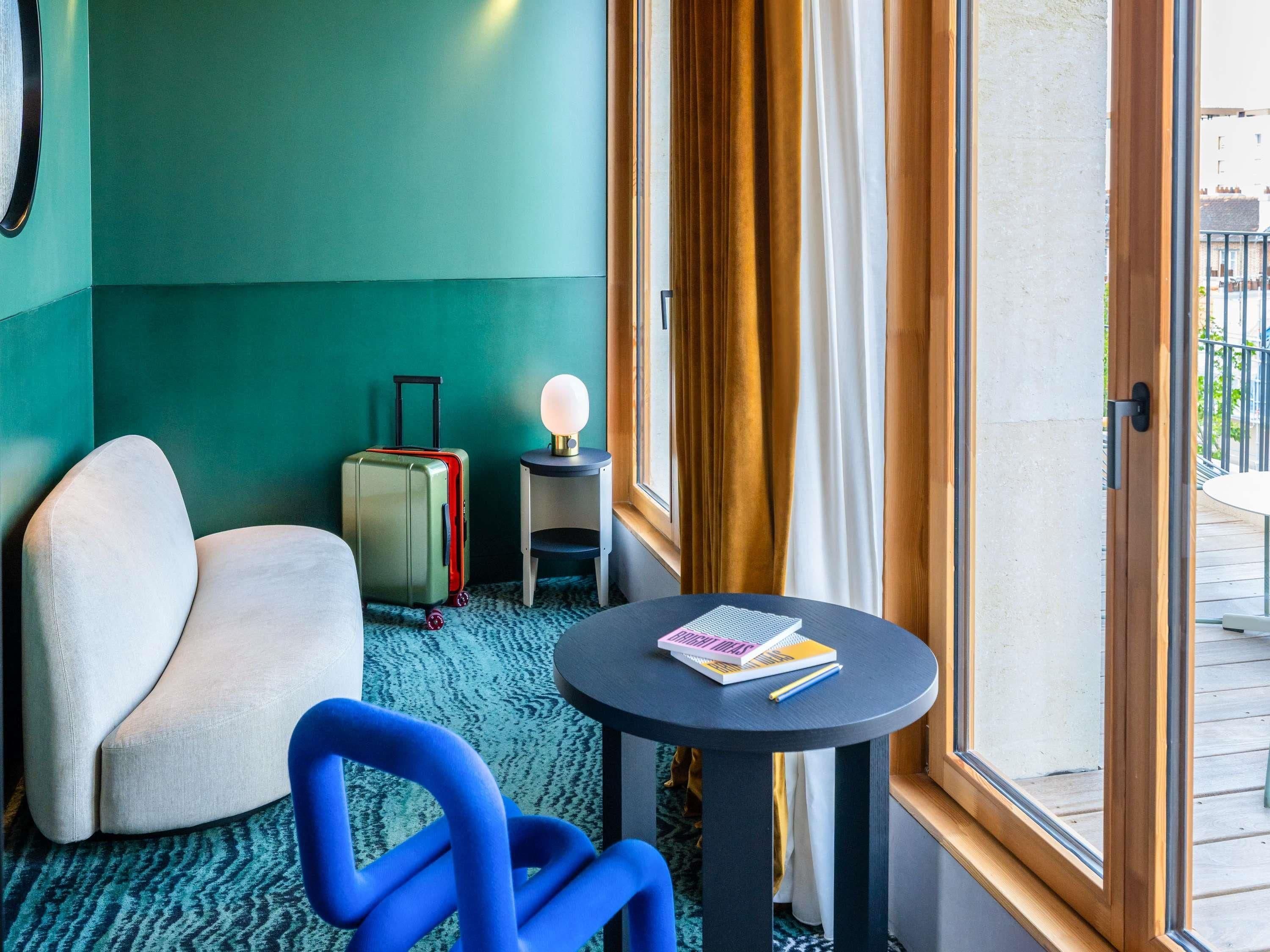 A hotel room with a green wall, a white armchair, a blue chair, a small table with a lamp, a suitcase, and a view of the outside balcony.
