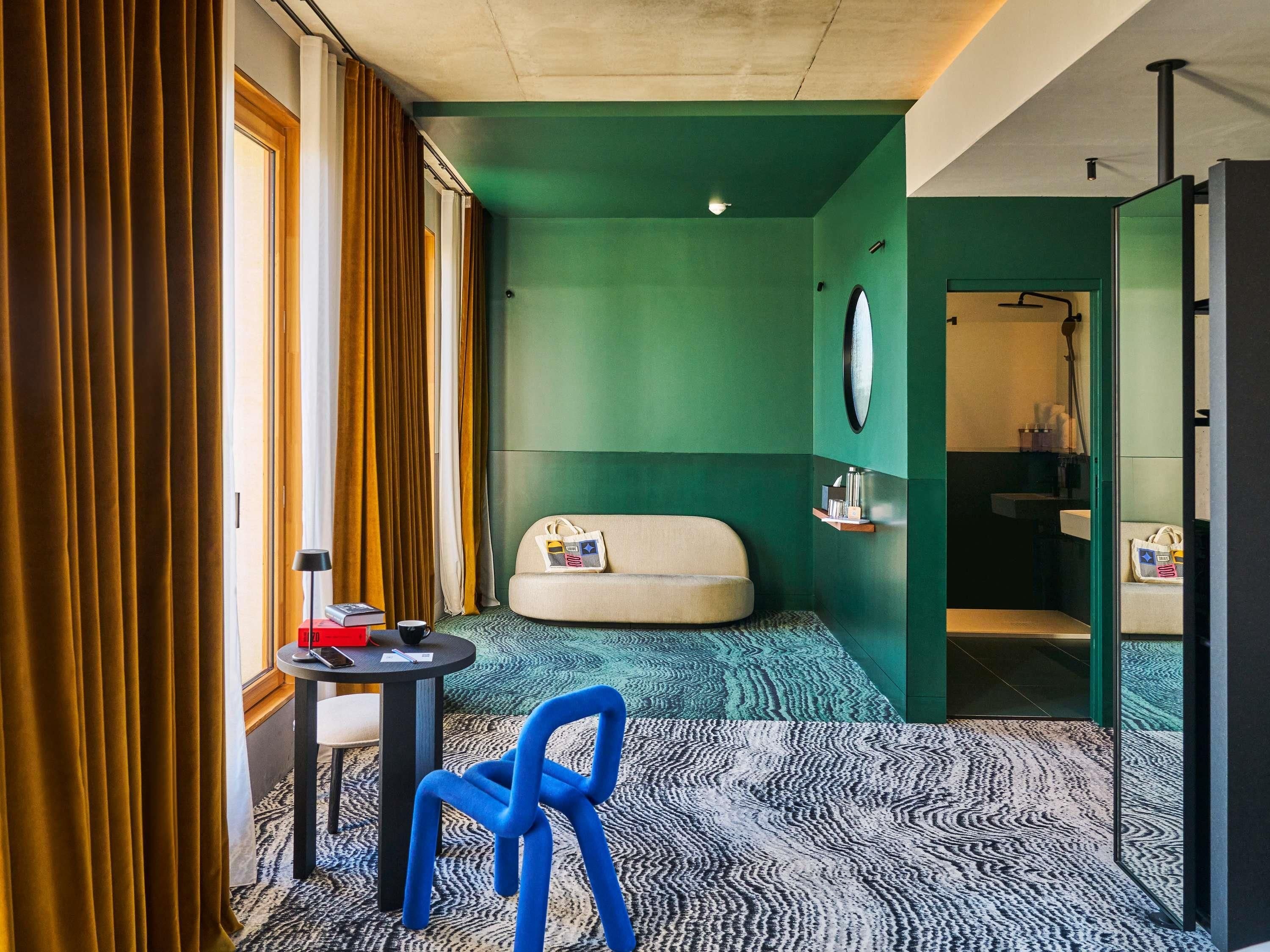 A hotel room with a sofa, a small table with a chair, a mirror, and a bathroom visible through an open door.