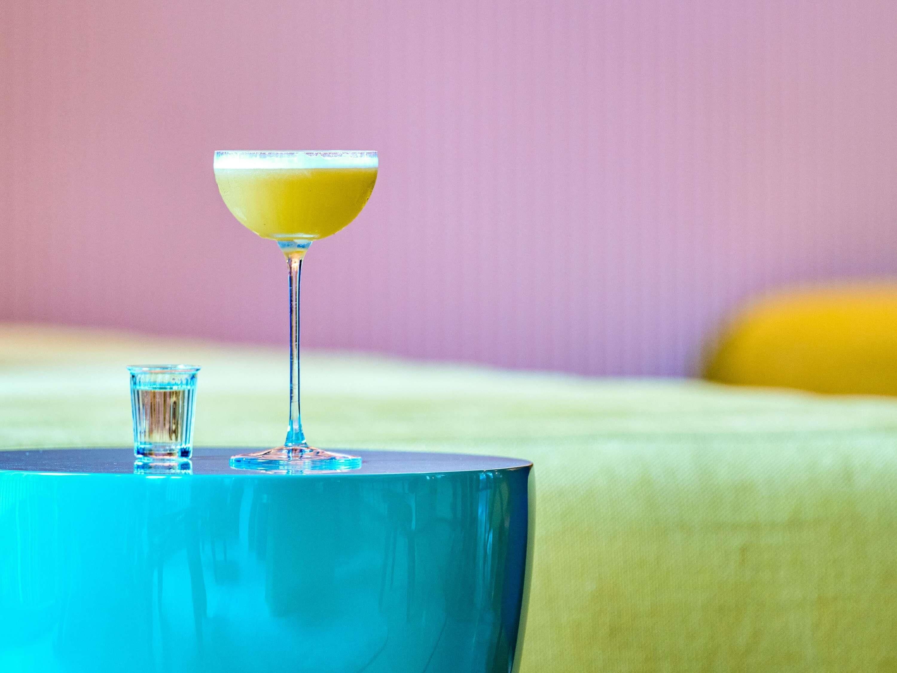 Um copo de cocktail sobre uma mesa azul, com uma cama ao fundo.