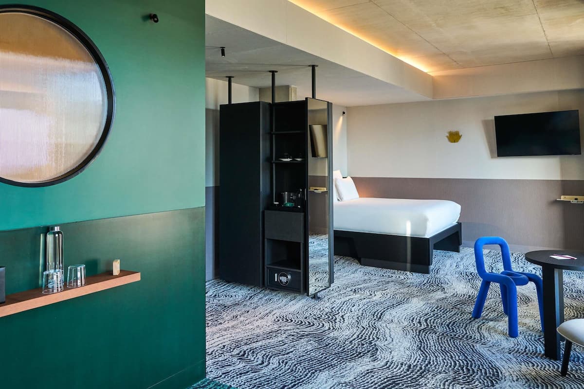 A hotel room with a bed, a TV, a shelf with glasses, and a patterned carpet.