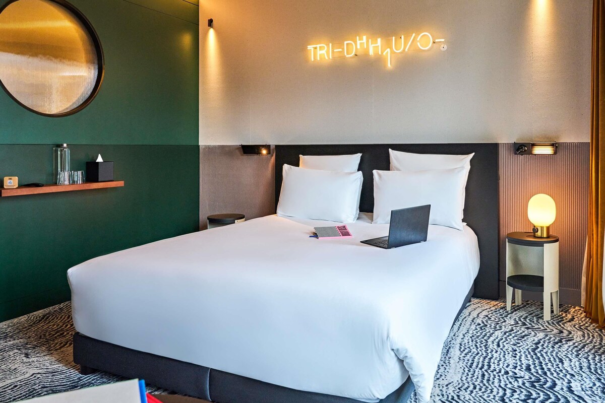 A modern hotel room with a neatly made bed, a bedside lamp, and a neon sign on the wall.