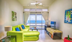 A bright living room with a green sofa, a TV, and a view of the ocean. - Vibrant and colorful ocean side villa with pool, AC and much more! (Akumal)