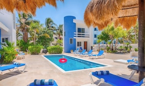 A pool area with blue lounge chairs, a red inflatable ring, and a building with blue and white exterior walls. - Vibrant and colorful ocean side villa with pool, AC and much more! (Akumal)