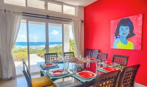 A dining area with a view of the ocean, a red wall with a painting, and a glass table set for four. - Vibrant and colorful ocean side villa with pool, AC and much more! (Akumal)