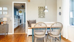 Uma área de refeições acolhedora com mesa de madeira e duas cadeiras, uma luminária pendente, e vista para a cozinha e sala de estar.