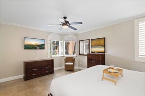 Une chambre à coucher avec un lit, un ventilateur de plafond, une commode, une chaise et des œuvres d’art accrochées aux murs.